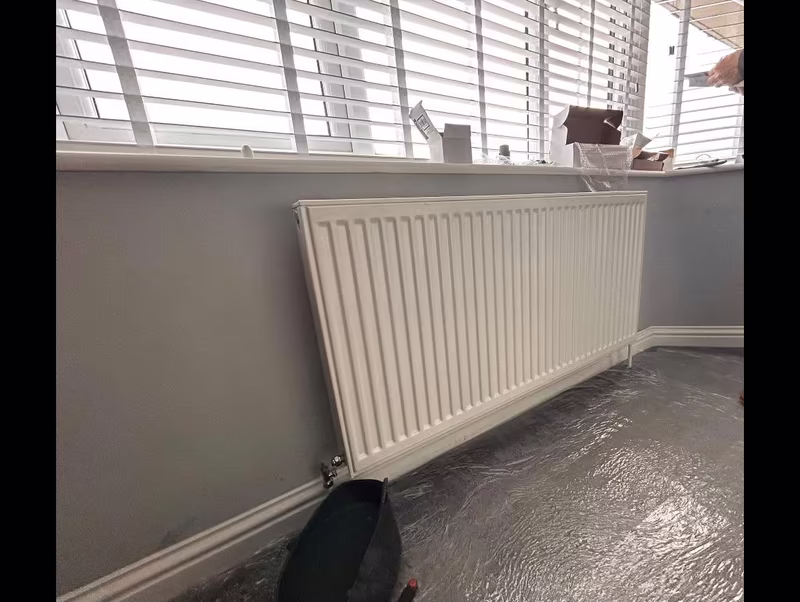 Old white panel radiator under a bay window, floor sheeted while the system is drained